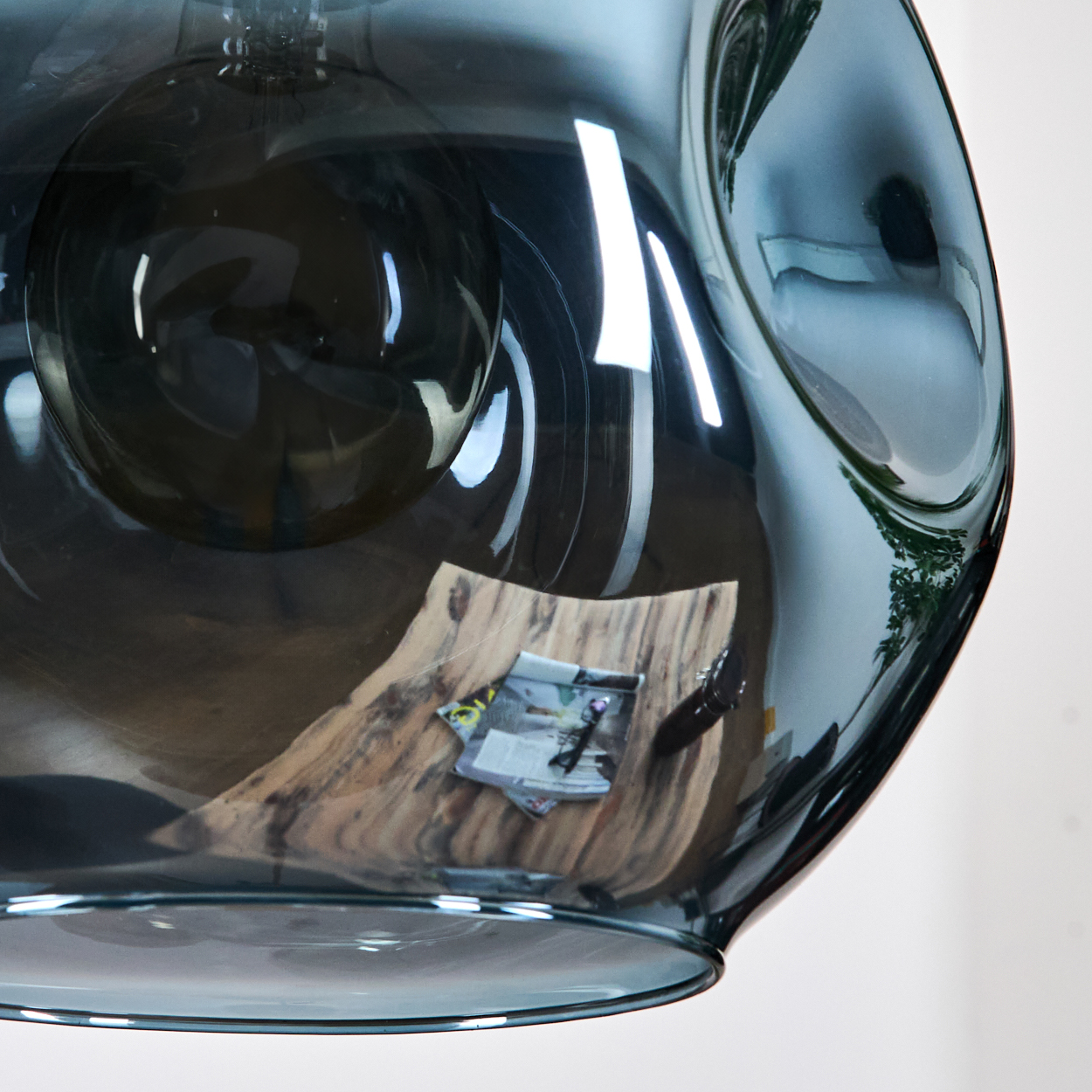 Ein dunkler Glasschirm reflektiert einen Holztisch mit Zeitschriften und Gegenständen. Der Hintergrund ist weiß.
