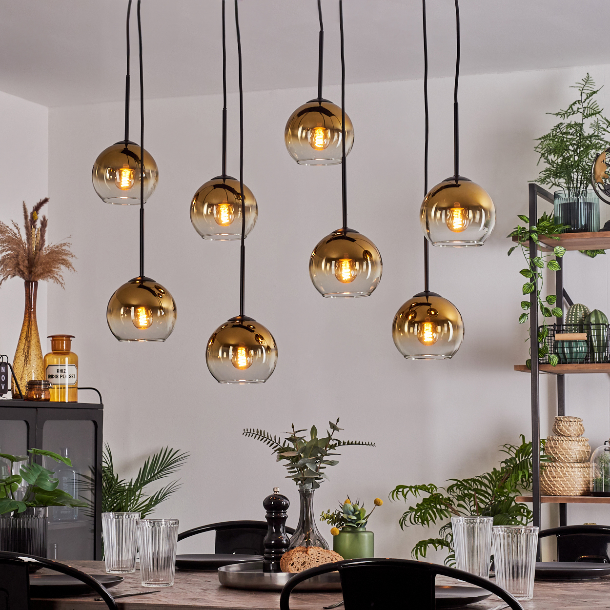 Esszimmer mit acht Hängelampen, Tisch mit Gläsern, Pflanzen und Regalen mit Dekorationen im Hintergrund.