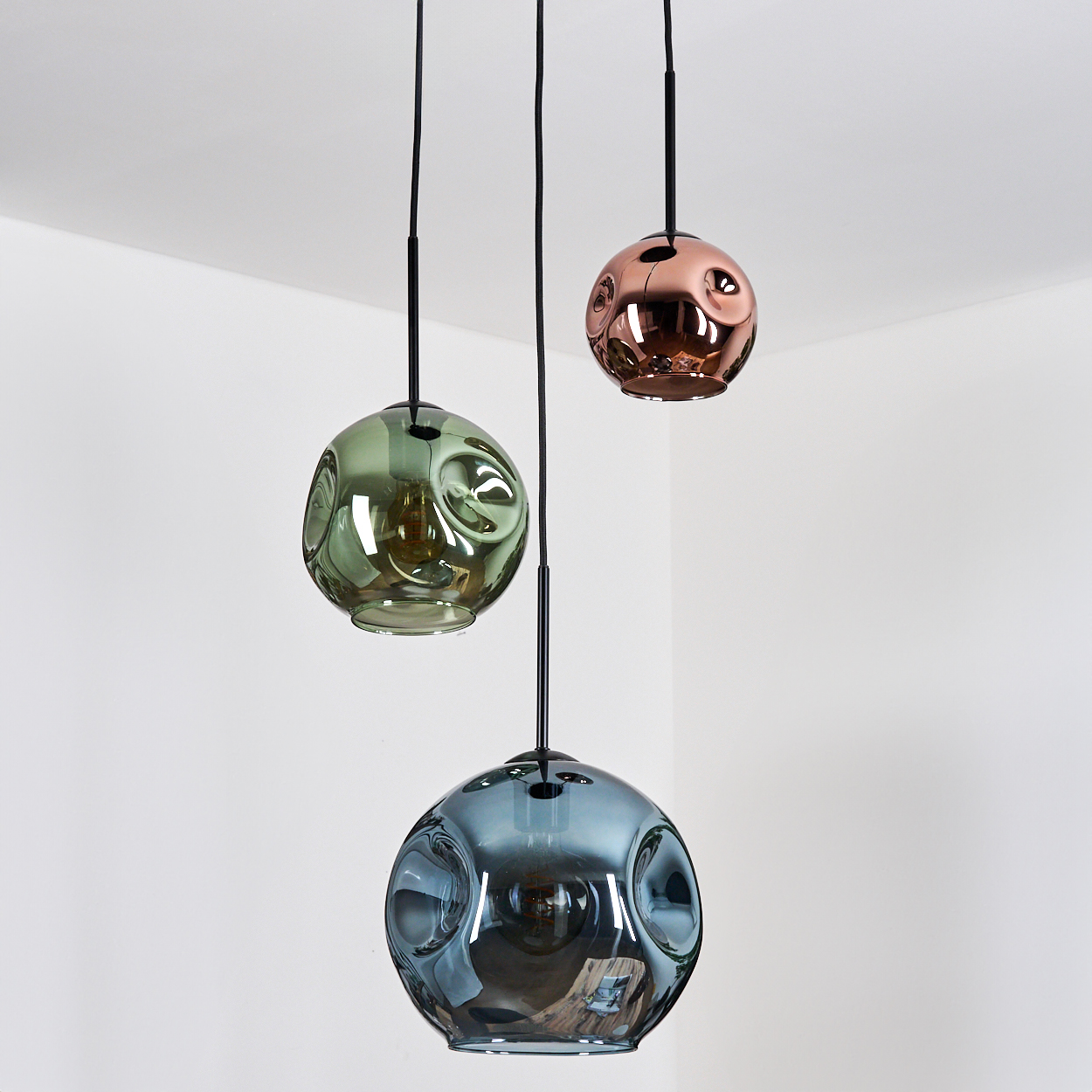 Drei kugelförmige Pendelleuchten hängen von der Decke. Eine ist kupferfarben, eine grün und eine blau. Der Hintergrund ist eine weiße Ecke.