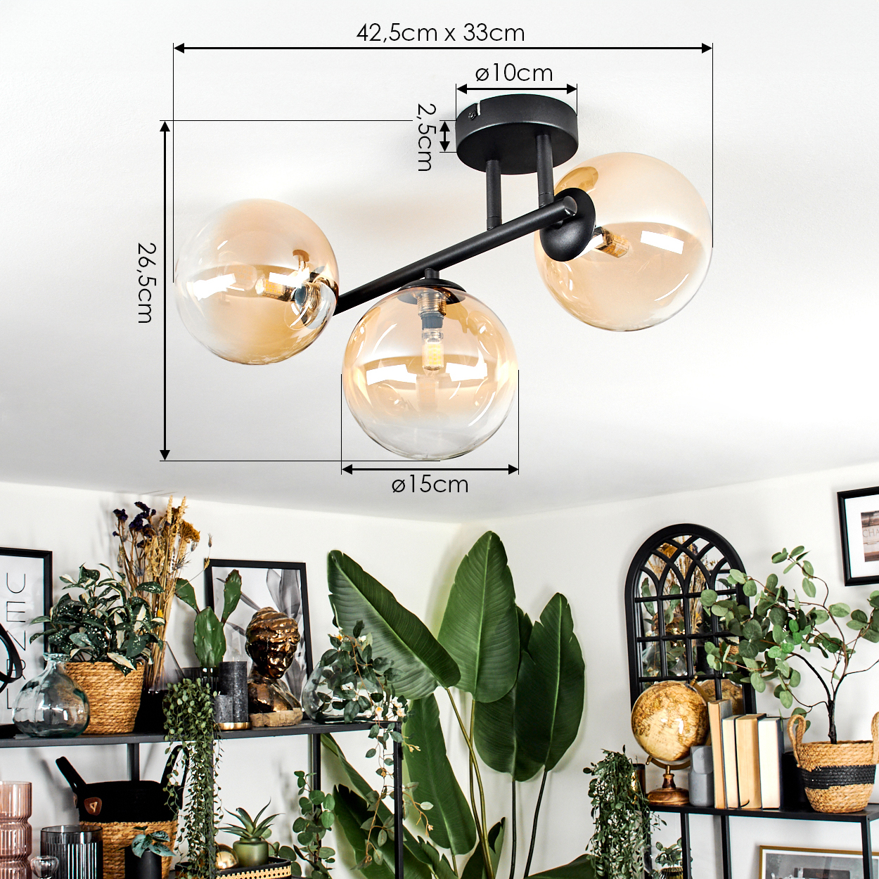 Eine moderne Deckenlampe mit drei bernsteinfarbenen Glaskugeln. Sie ist auf einem schwarzen Rahmen montiert. Im Hintergrund sind verschiedene Pflanzen und Dekorationsartikel zu sehen.