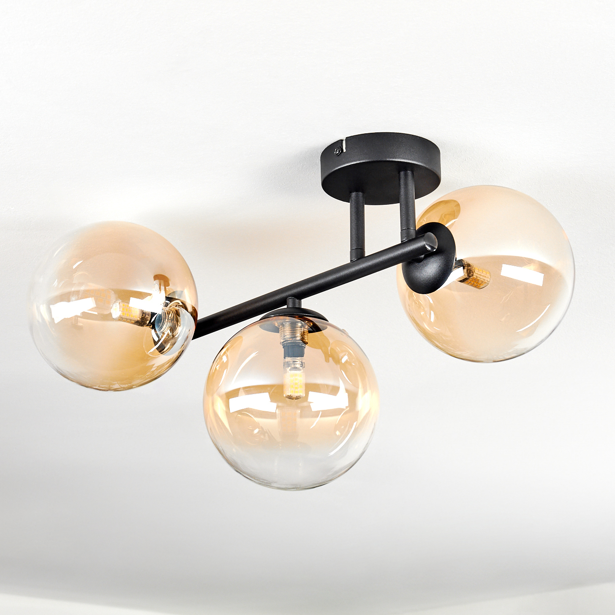 Moderne Deckenleuchte mit drei bernsteinfarbenen Glaskugeln. Schwarze Metallarme und Deckenhalterung. Weißer Deckenhintergrund.