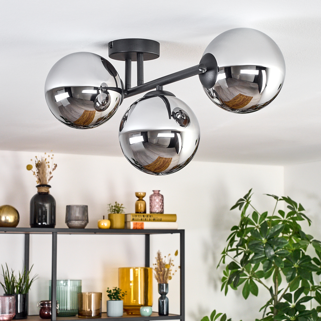Moderne Deckenlampe mit drei Chromkugeln. Darunter ein Regal mit Vasen, Pflanzen und Büchern. Rechts eine große grüne Pflanze.