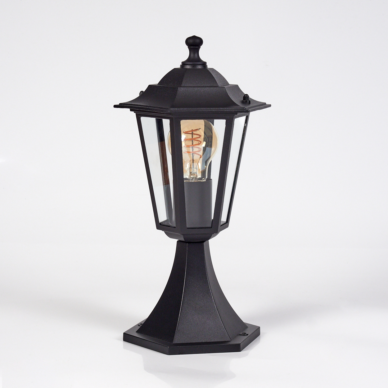 Eine schwarze Außenlampe mit Glasgehäuse. Sie hat eine Glühbirne im Inneren. Die Lampe steht auf einer weißen Oberfläche.