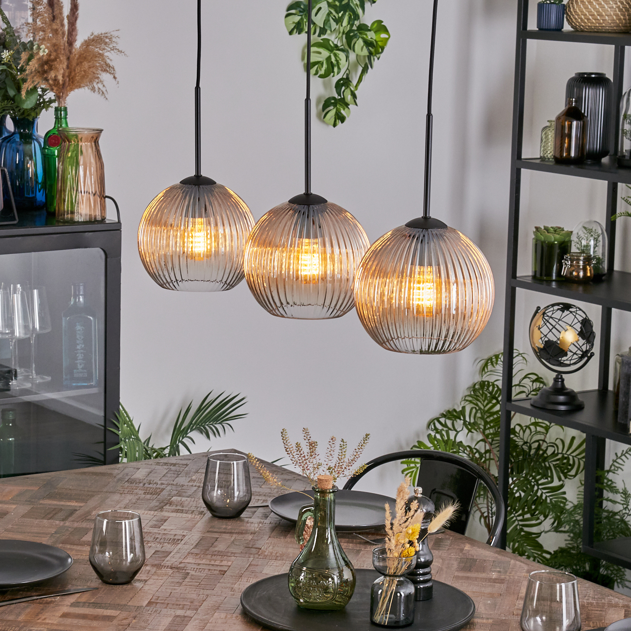Drei Pendelleuchten über einem Holztisch mit Gläsern und Dekoration. Regale mit Vasen und einem Globus sind im Hintergrund.
