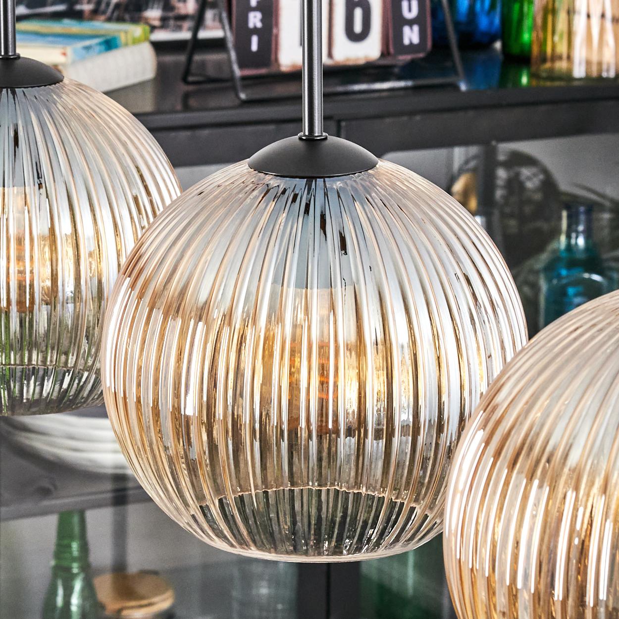 Drei Glas-Pendelleuchten mit geripptem Design. Die Leuchten haben einen warmen, bernsteinfarbenen Schein. Hintergrund zeigt ein Regal mit Flaschen.