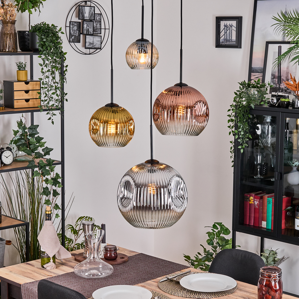 Esszimmer mit vier Glas-Pendelleuchten. Holztisch mit Tellern und Dekanter. Pflanzen und Regale im Hintergrund.