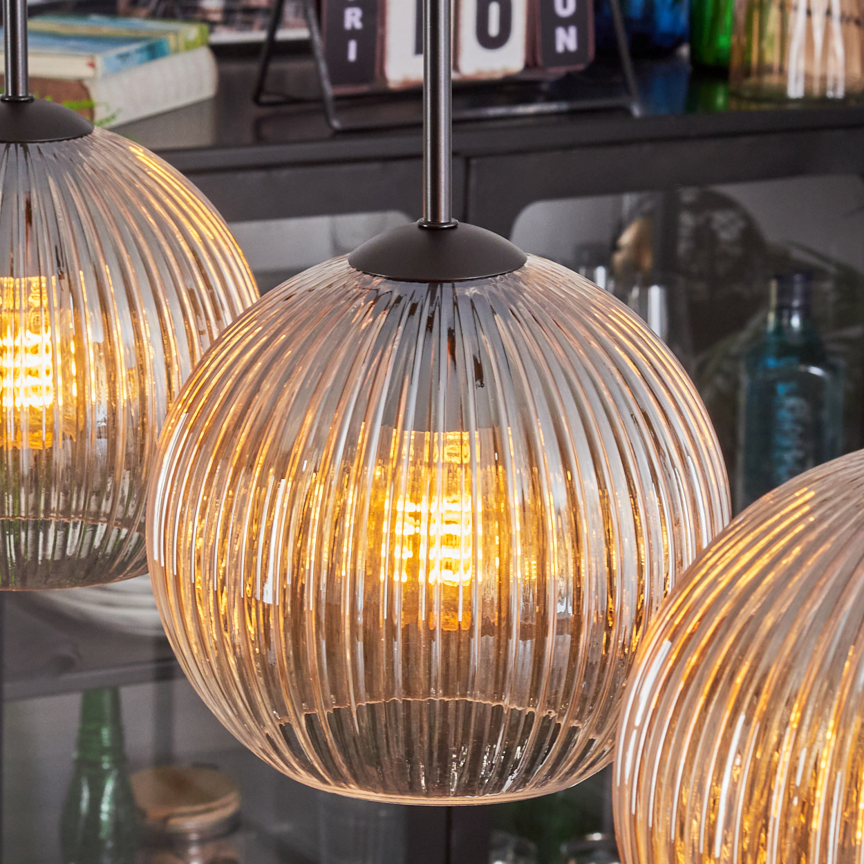 Drei Pendelleuchten mit geriffelten Glasschirmen, die mit warmem Licht beleuchtet werden. Im Hintergrund Regale mit Flaschen und Gegenständen.