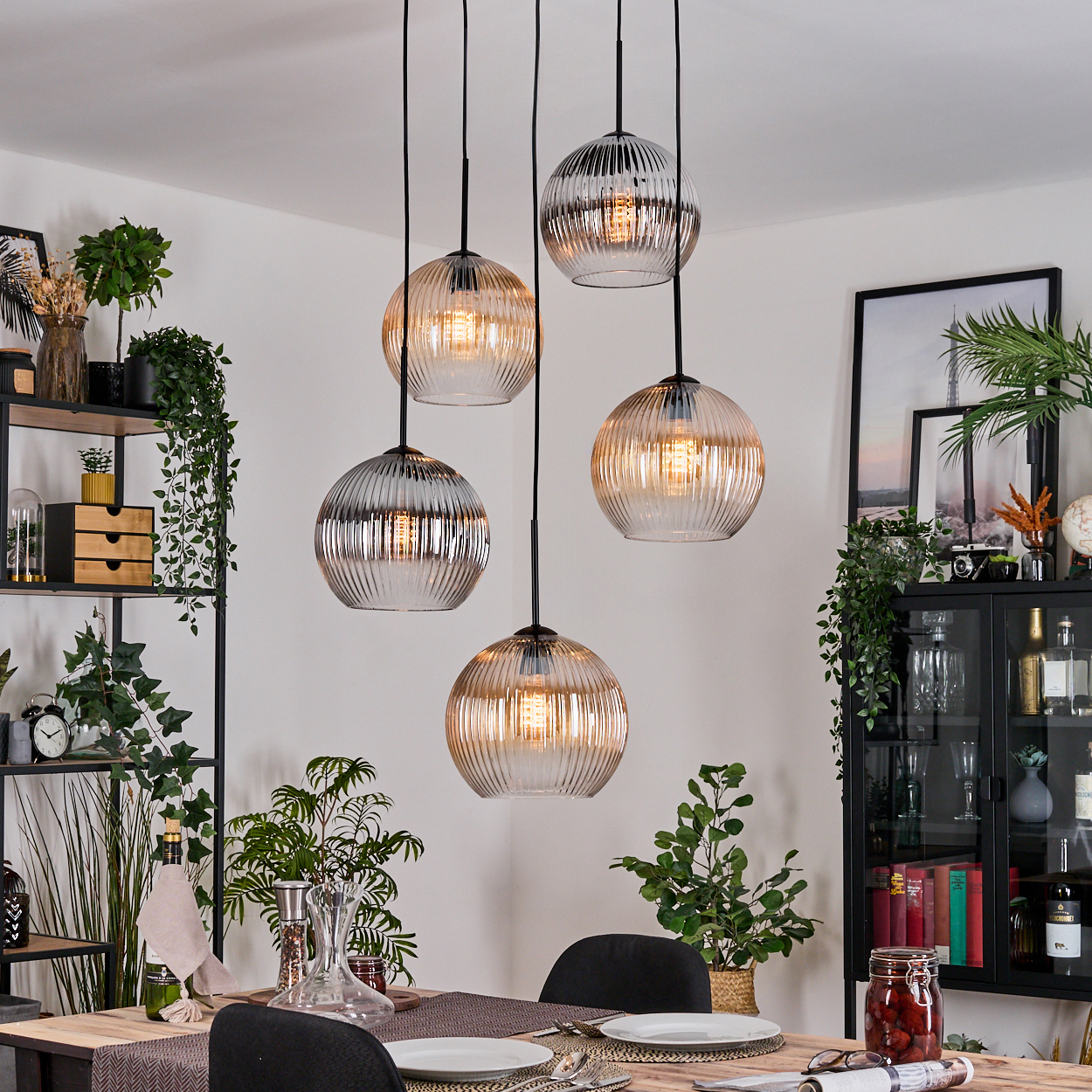 Esszimmer mit Hängelampen. Ein Tisch ist mit Tellern und Besteck gedeckt. Regale und ein Schrank sind mit Pflanzen und Dekoration zu sehen.