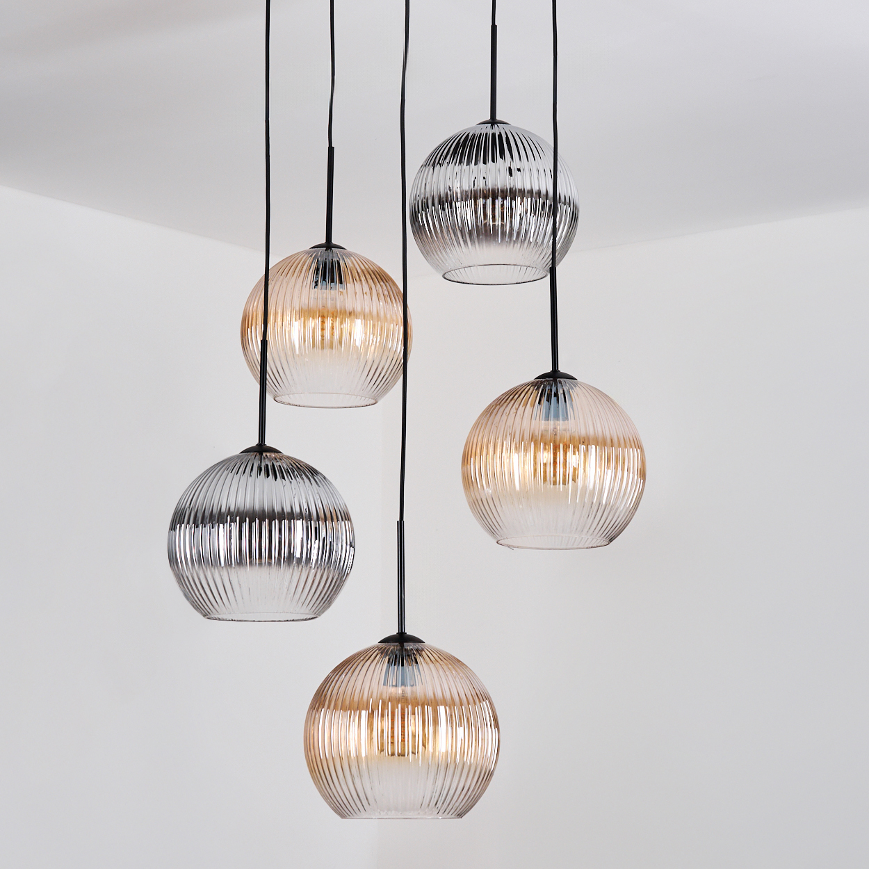 Fünf Pendelleuchten aus Glas hängen von der Decke. Einige sind silberfarben, andere bernsteinfarben, alle mit schwarzen Kabeln an einer weißen Wand.