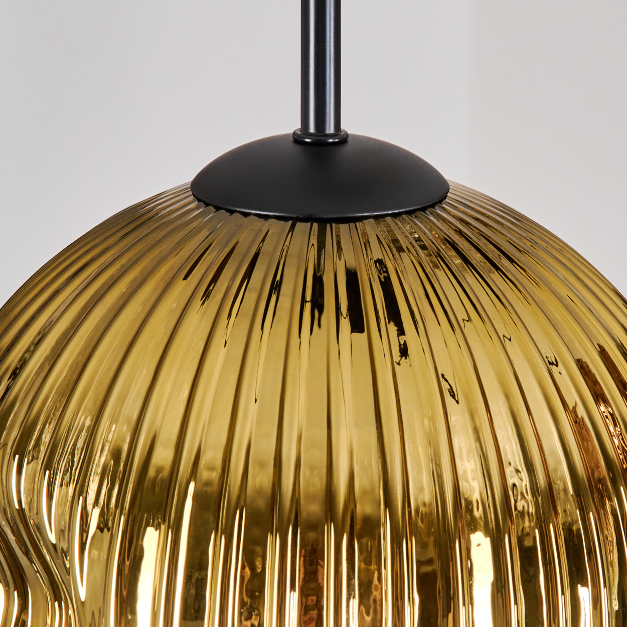 Nahaufnahme einer hängenden goldfarbenen Lampe. Sie hat einen schwarzen Deckel und einen gerippten, kugelförmigen Schirm. Der Hintergrund ist weiß.