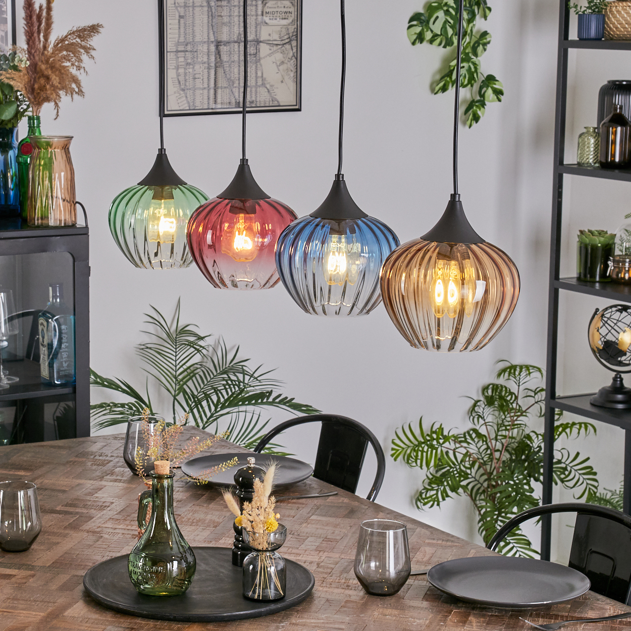Vier bunte Pendelleuchten hängen über einem Holztisch, der zum Abendessen gedeckt ist. Pflanzen und dekorative Gegenstände befinden sich im Hintergrund.