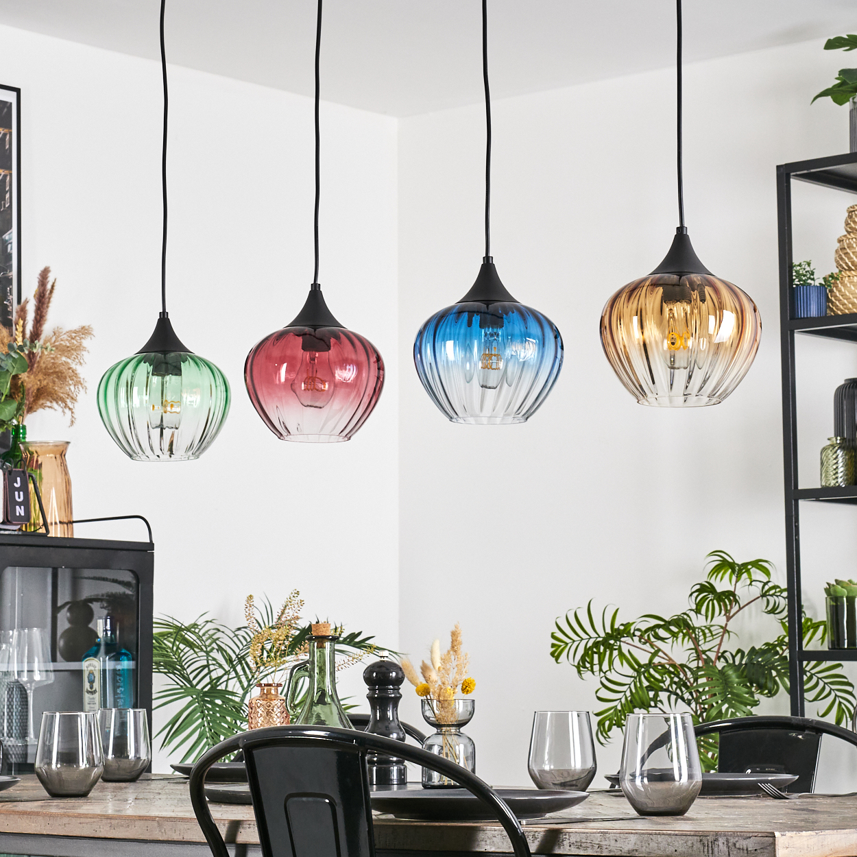 Vier bunte Pendelleuchten über einem Holztisch, der für das Abendessen gedeckt ist. Pflanzen und Dekoration sind ebenfalls vorhanden.
