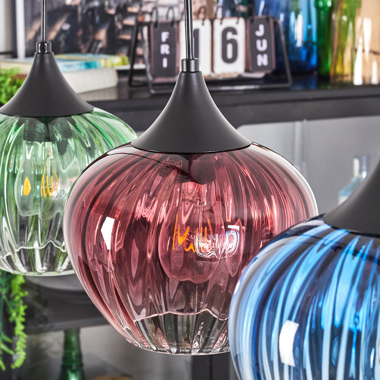Drei bunte Glas-Pendelleuchten hängen. Die mittlere ist rot, flankiert von grün und blau. Im Hintergrund sind ein Kalender und andere Objekte zu sehen.