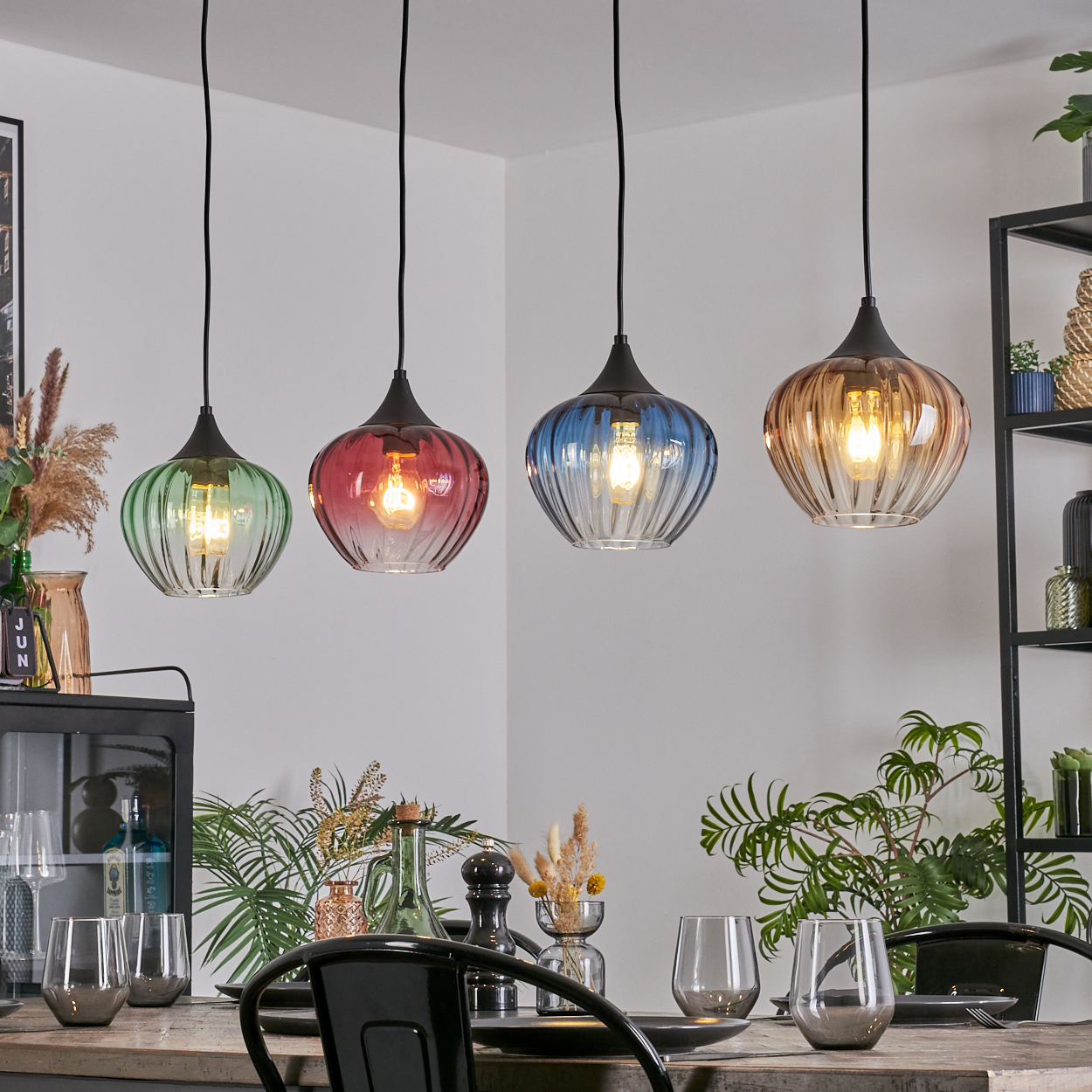 Vier bunte Pendelleuchten über einem Holztisch, der zum Abendessen gedeckt ist. Pflanzen, Gläser und dekorative Elemente sind vorhanden.