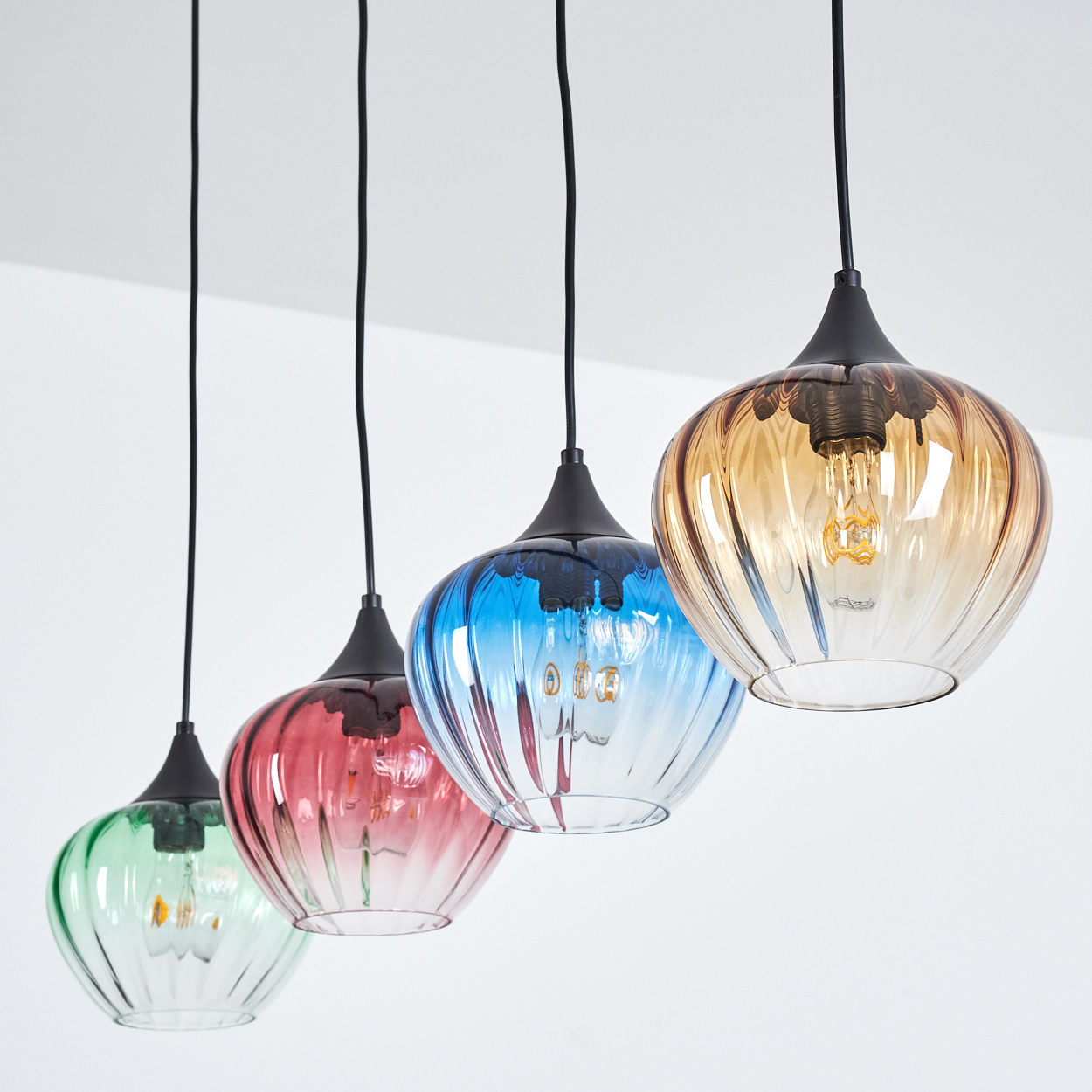 Vier bunte Pendelleuchten hängen von einer weißen Decke. Jede hat einen andersfarbigen Glasschirm: grün, rot, blau und bernsteinfarben.
