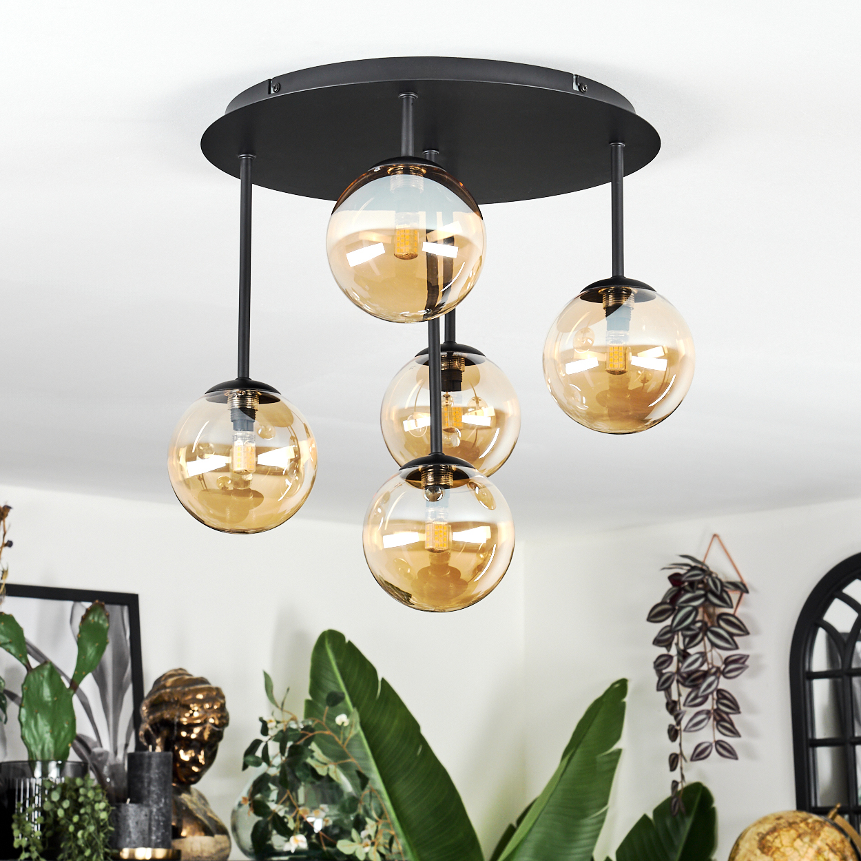 Eine moderne Deckenlampe mit fünf Glaskugeln, schwarze Halterung. Pflanzen in Töpfen und eine Büste sind im Hintergrund zu sehen.