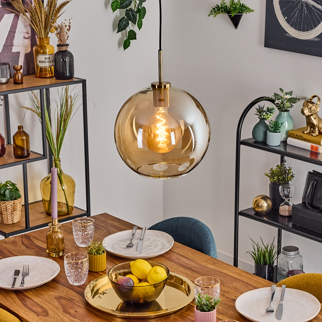 Ein Esstisch mit goldener Pendelleuchte, gedeckt mit Tellern, Gläsern und einer Schüssel mit Zitronen. Regale mit Dekoration im Hintergrund.