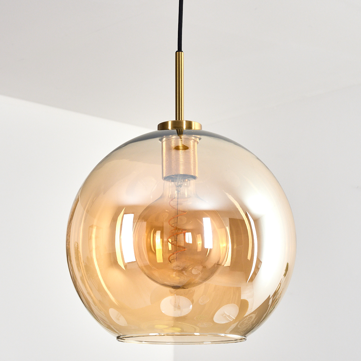 Eine runde, bernsteinfarbene Glashängeleuchte mit sichtbarer Glühbirne hängt von der Decke. Goldene Akzente und ein schwarzes Kabel sind sichtbar.