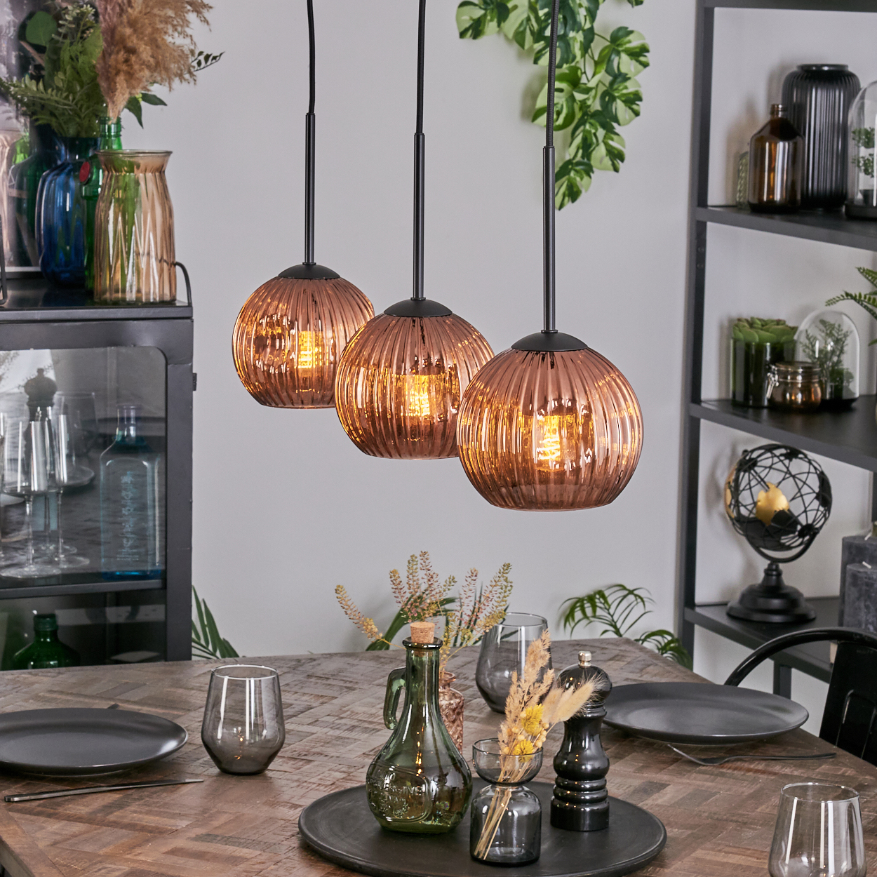 Drei kupferfarbene Pendelleuchten hängen über einem Holztisch, der mit Tellern, Gläsern und Dekorationsartikeln gedeckt ist. Regale halten Vasen und Pflanzen.