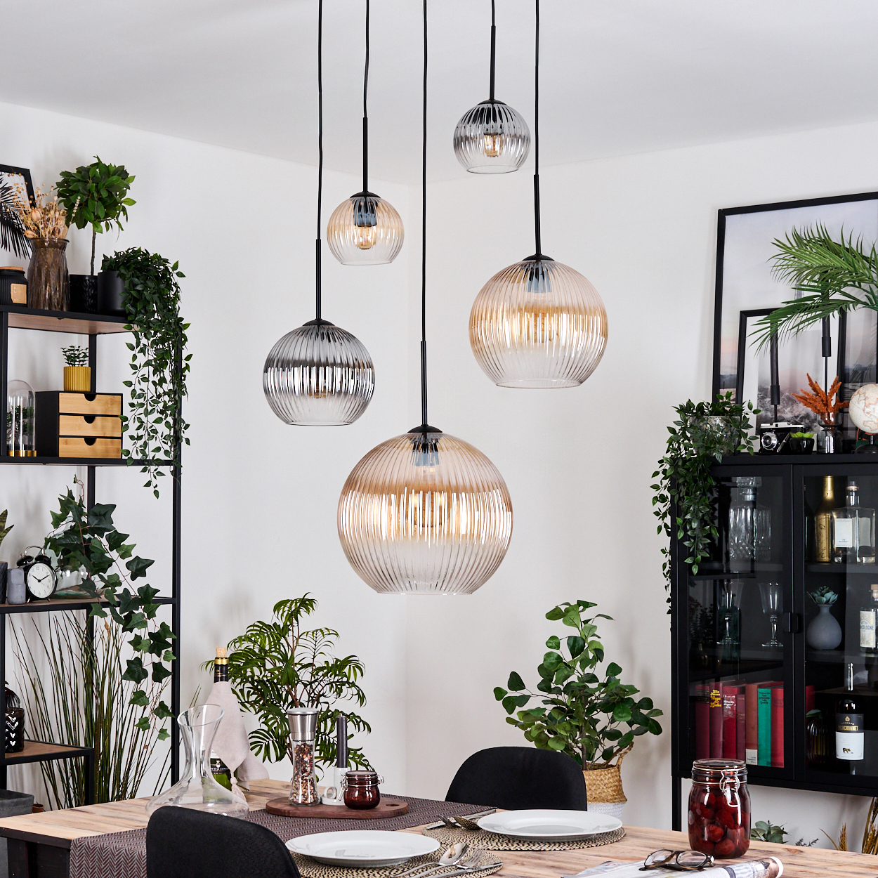 Esszimmer mit hängenden Kugelleuchten. Tisch mit Tellern und Besteck. Pflanzen und ein schwarzer Schrank mit Dekoration im Hintergrund.