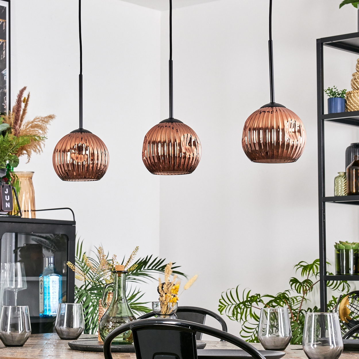 Drei kupferfarbene Pendelleuchten über einem Esstisch mit Gläsern, Pflanzen und einem schwarzen Metallregal im Hintergrund.