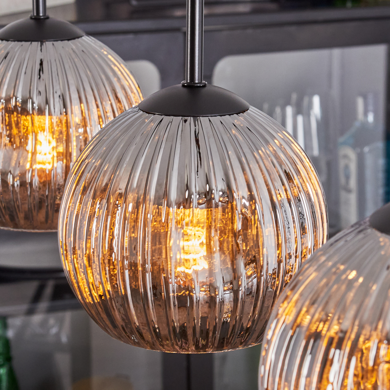 Drei Glas-Hängeleuchten mit strukturierter Oberfläche, die Licht reflektieren. Im Hintergrund ein verschwommenes Interieur mit dunklen Elementen.