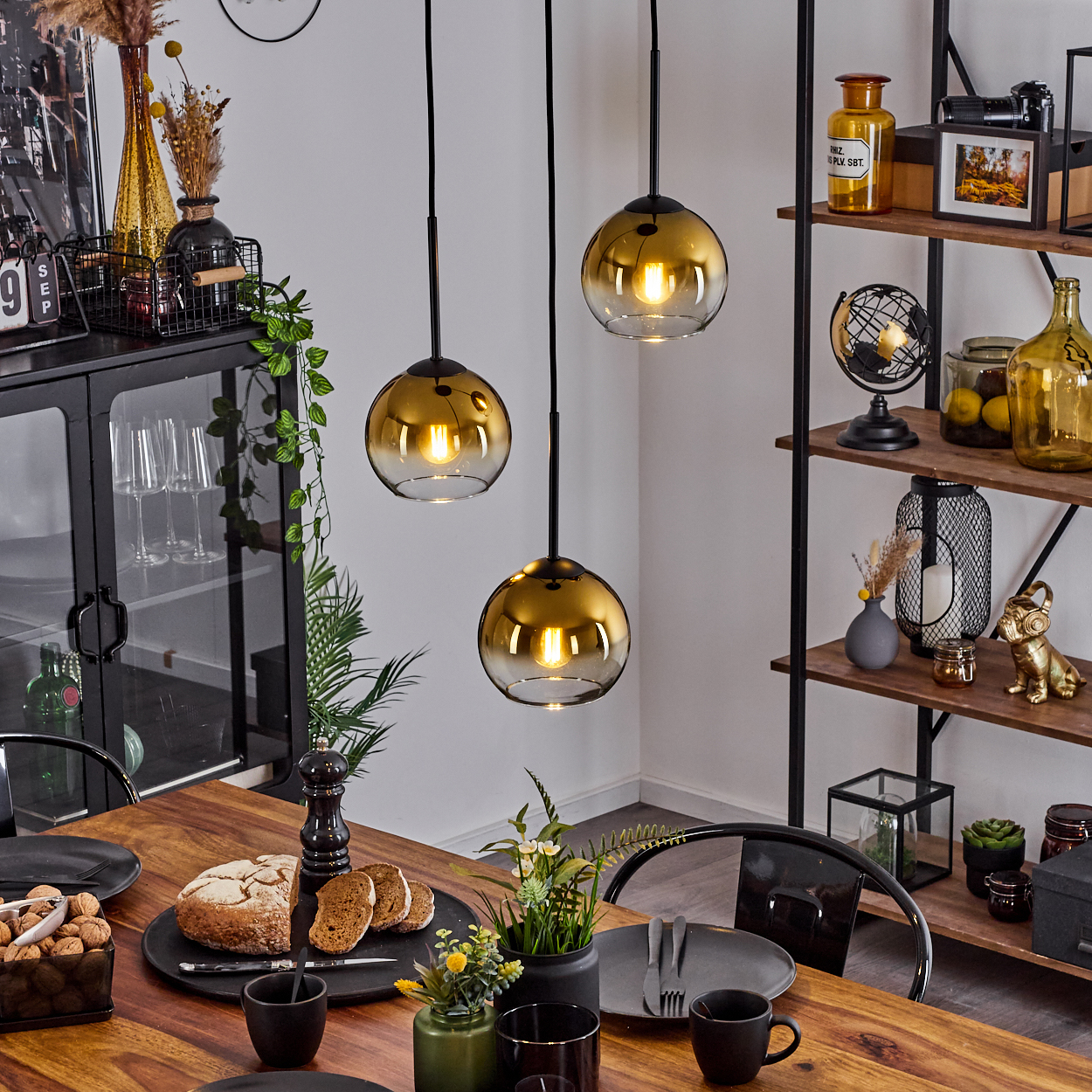 Esszimmer mit Holztisch, drei Hängelampen, schwarzem Schrank, Regal mit Dekorationen und Brot mit Scheiben.