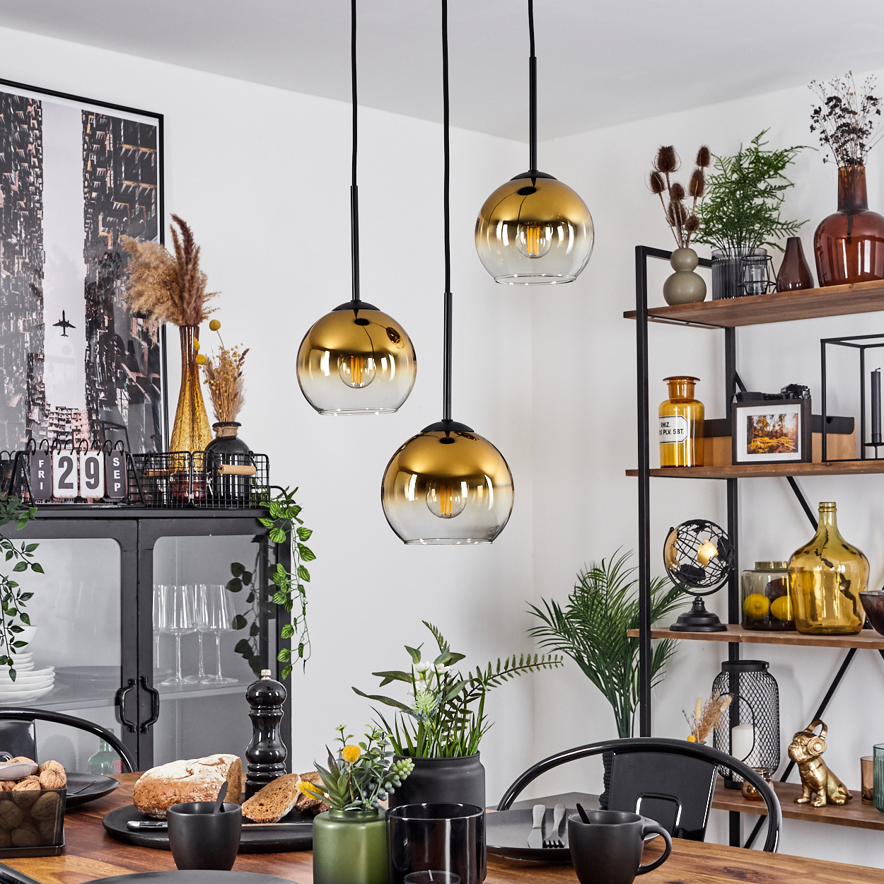 Esszimmer mit drei goldenen Pendelleuchten, Tisch mit Speisen und Getränken sowie ein schwarzes Regal mit Dekorationen.