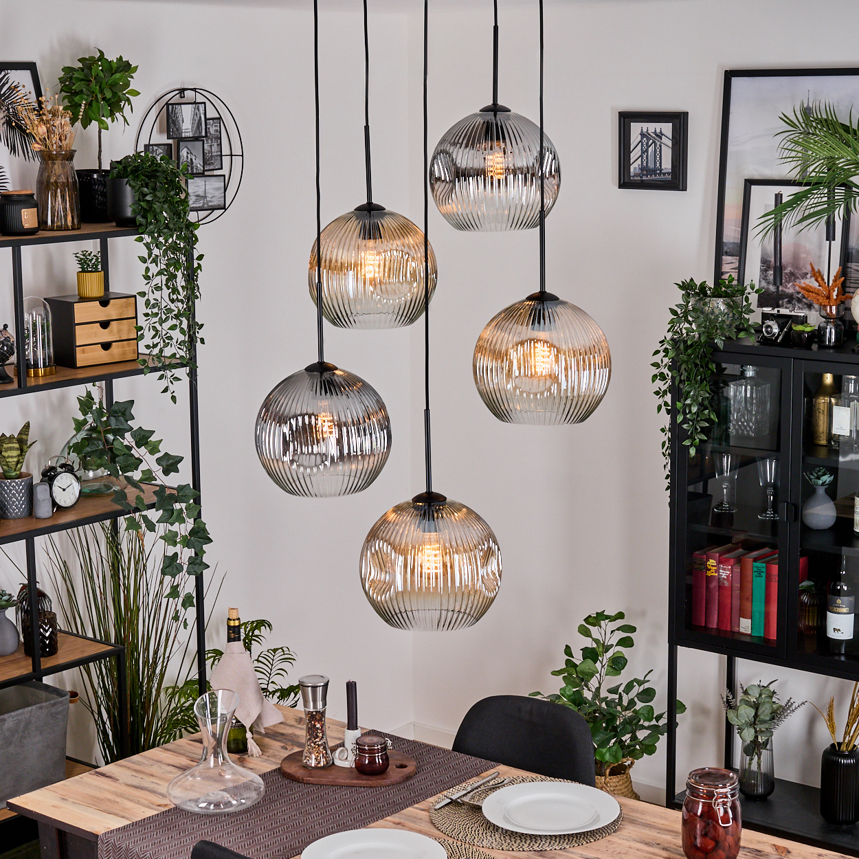 Esszimmer mit Holztisch, Hängelampen, Regalen mit Pflanzen und einem Schrank mit Gläsern und Flaschen.
