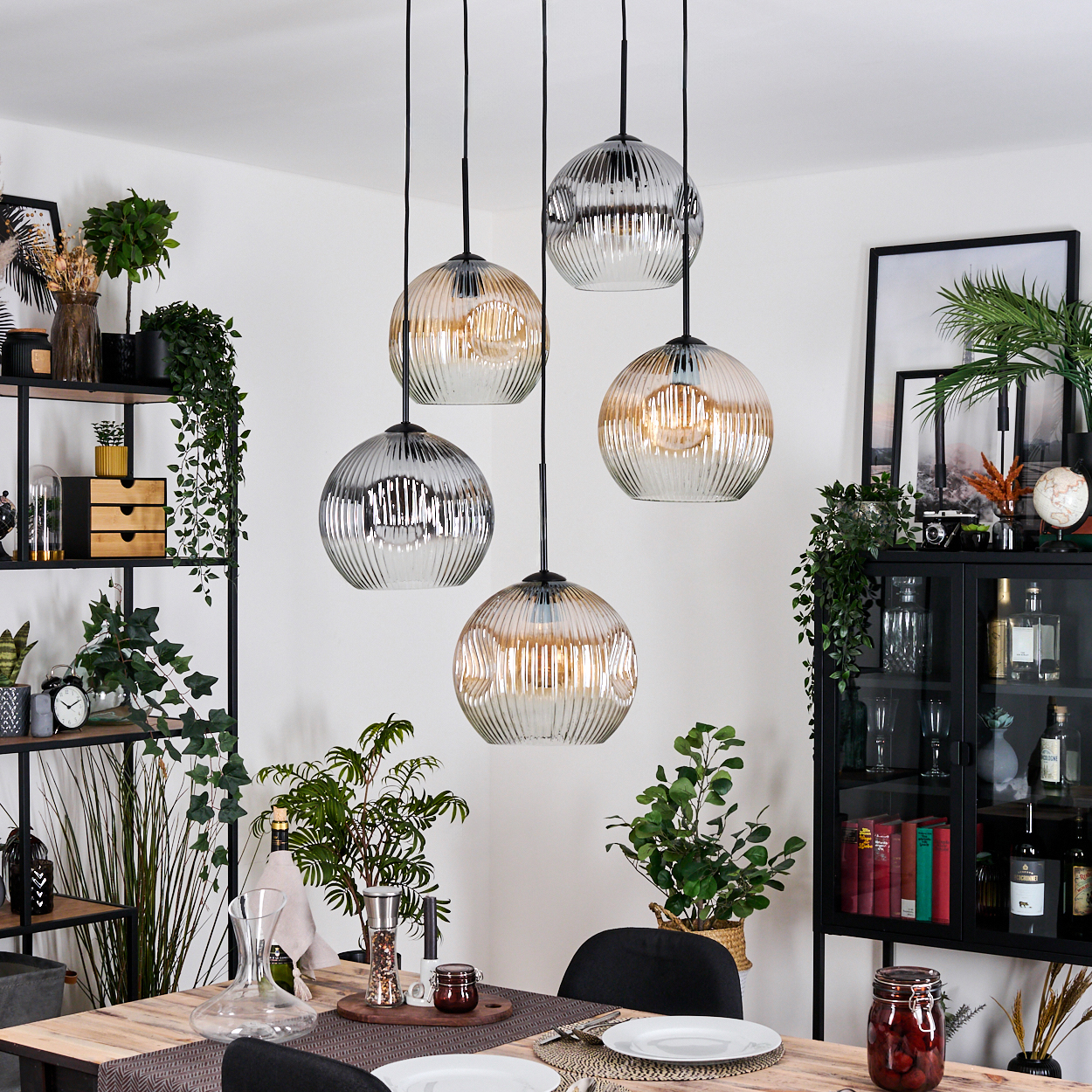 Esszimmer mit Holztisch, Hängelampen, Pflanzen und einem schwarzen Schrank mit Dekoration. Der Raum ist gut beleuchtet.