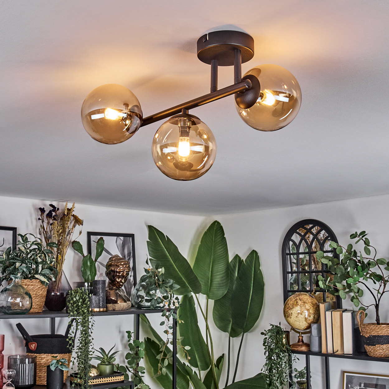 Eine moderne Deckenleuchte mit drei bernsteinfarbenen Kugeln beleuchtet einen Raum voller Pflanzen, Regale und Kunstwerke.