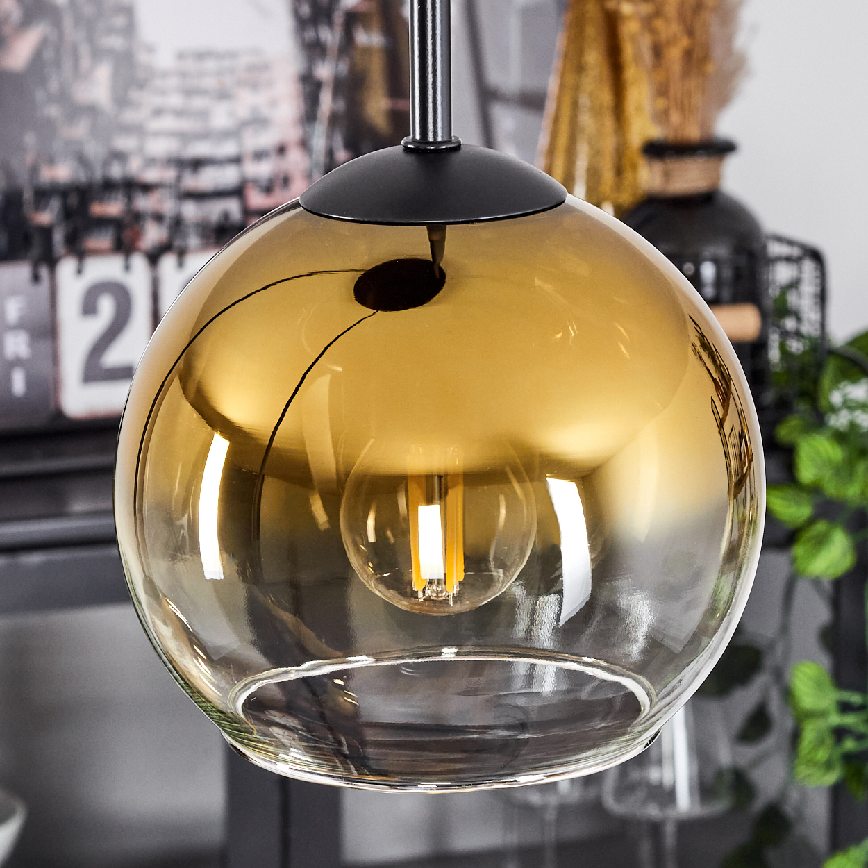 Eine goldene Glaskugel-Pendelleuchte mit einer leuchtenden Glühbirne im Inneren. Im Hintergrund befinden sich ein Kalender, Pflanzen und eine Vase.