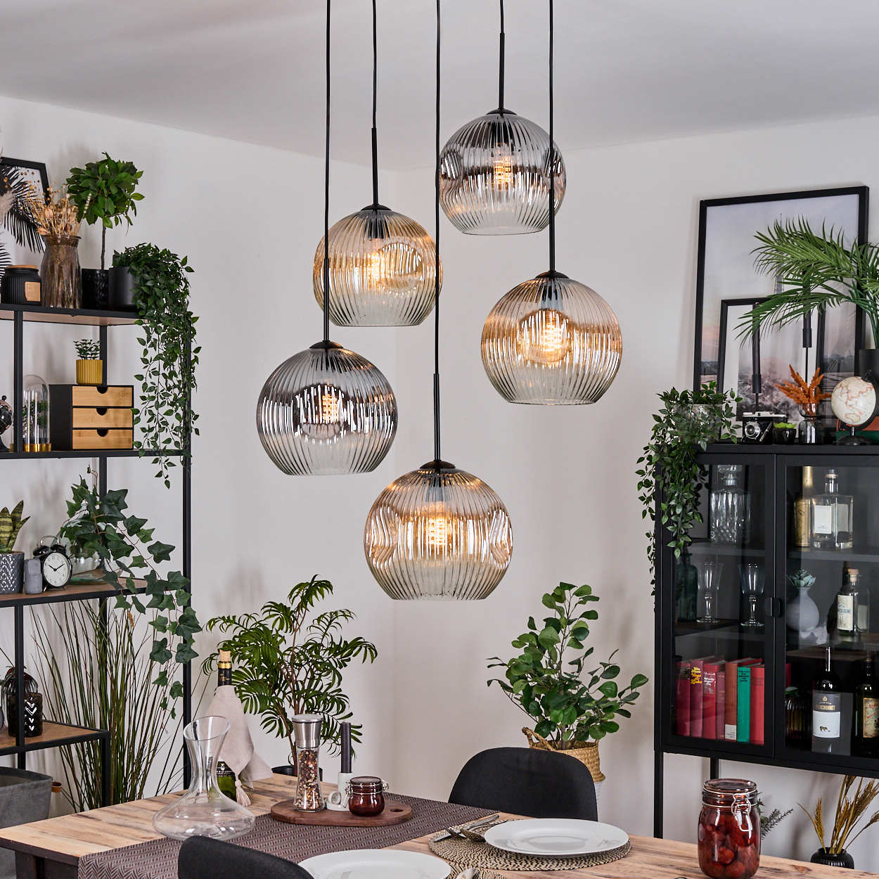 Esszimmer mit hängenden Kugelleuchten, einem Holztisch, Pflanzen und einem schwarzen Schrank. Die Szene ist hell und modern.