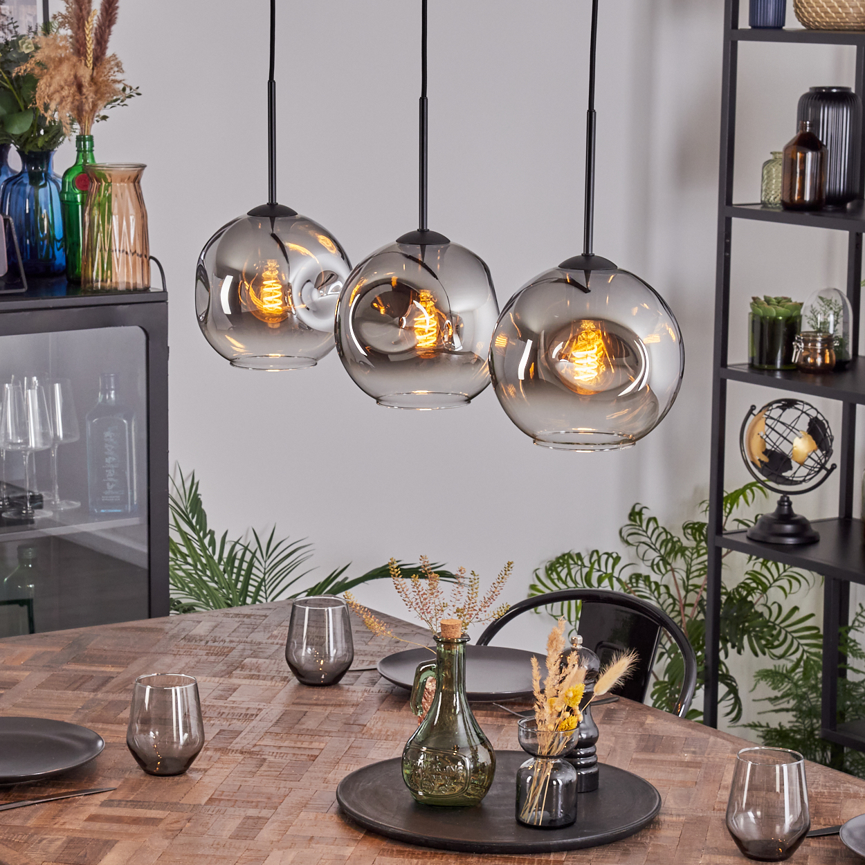 Drei Hängelampen hängen über einem Holztisch, der für das Abendessen gedeckt ist. Vasen und ein schwarzes Regal sind im Hintergrund.