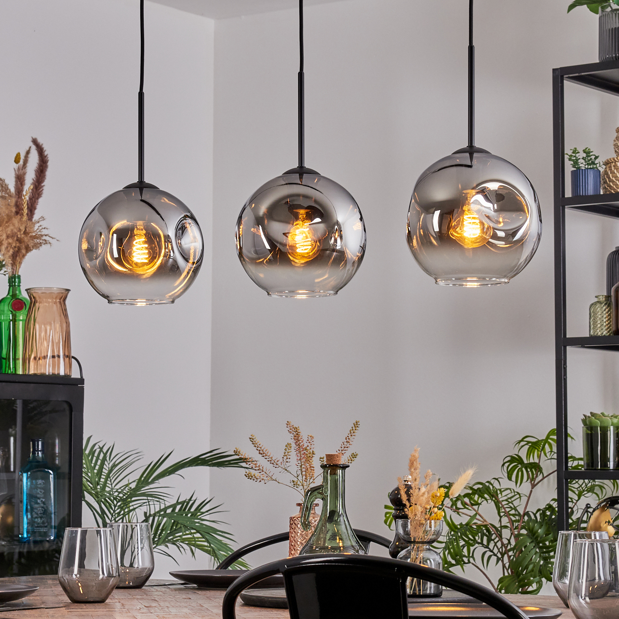 Drei Pendelleuchten mit warmen Glühbirnen hängen über einem Tisch, der für das Abendessen gedeckt ist. Pflanzen, Gläser und ein Metallregal sind im Hintergrund.