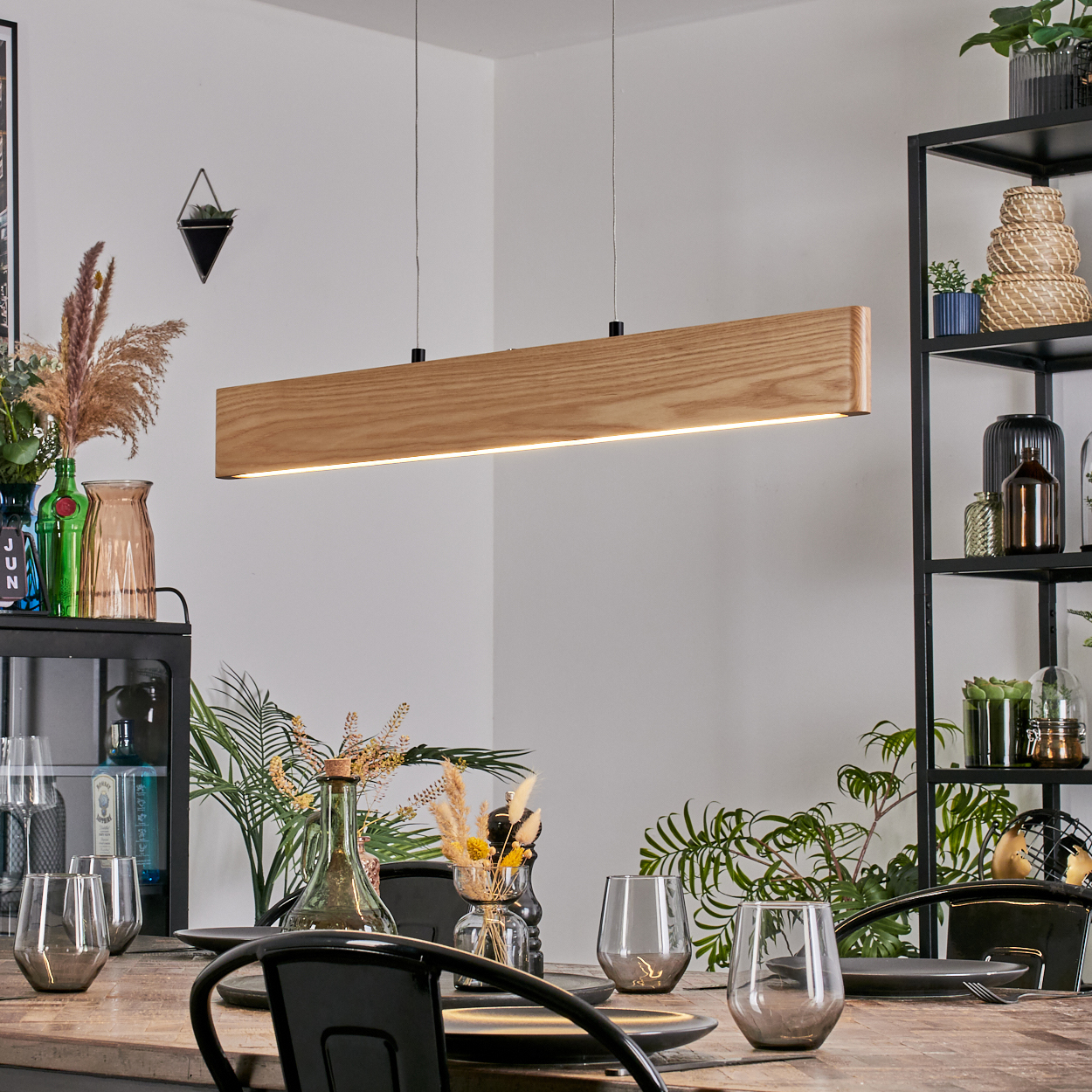Esszimmer mit rechteckiger Holzleuchte. Tisch mit Gläsern und Tellern. Regale mit Deko im Hintergrund.