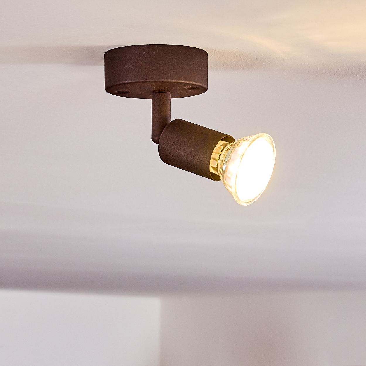 Eine braune Scheinwerferbefestigung ist an einer weißen Decke montiert. Das Licht ist an und beleuchtet den Raum darunter. Die Halterung ist abgewinkelt.