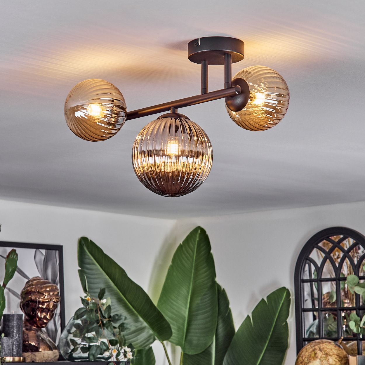 Eine Deckenleuchte mit drei Glasschirmen über grünen Pflanzen und Dekorationsartikeln. Die Lichter sind an.