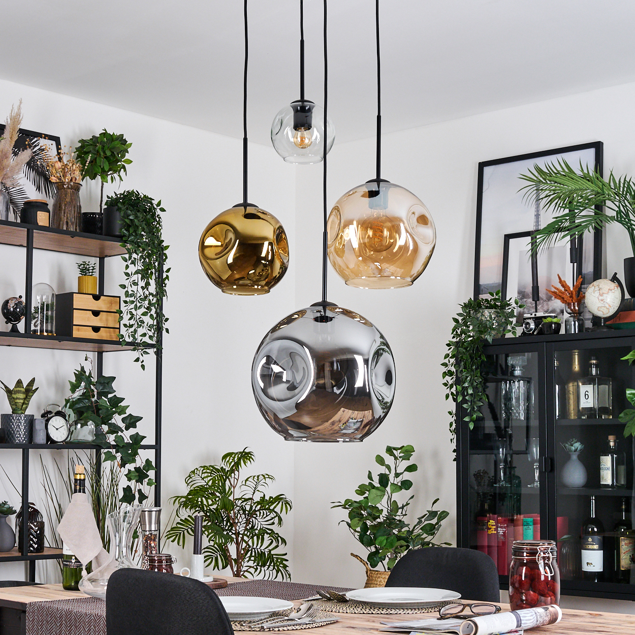 Esszimmer mit drei Hängelampen. Ein Tisch ist mit Tellern und Gläsern gedeckt. Regale und ein Schrank halten Pflanzen und Dekor.