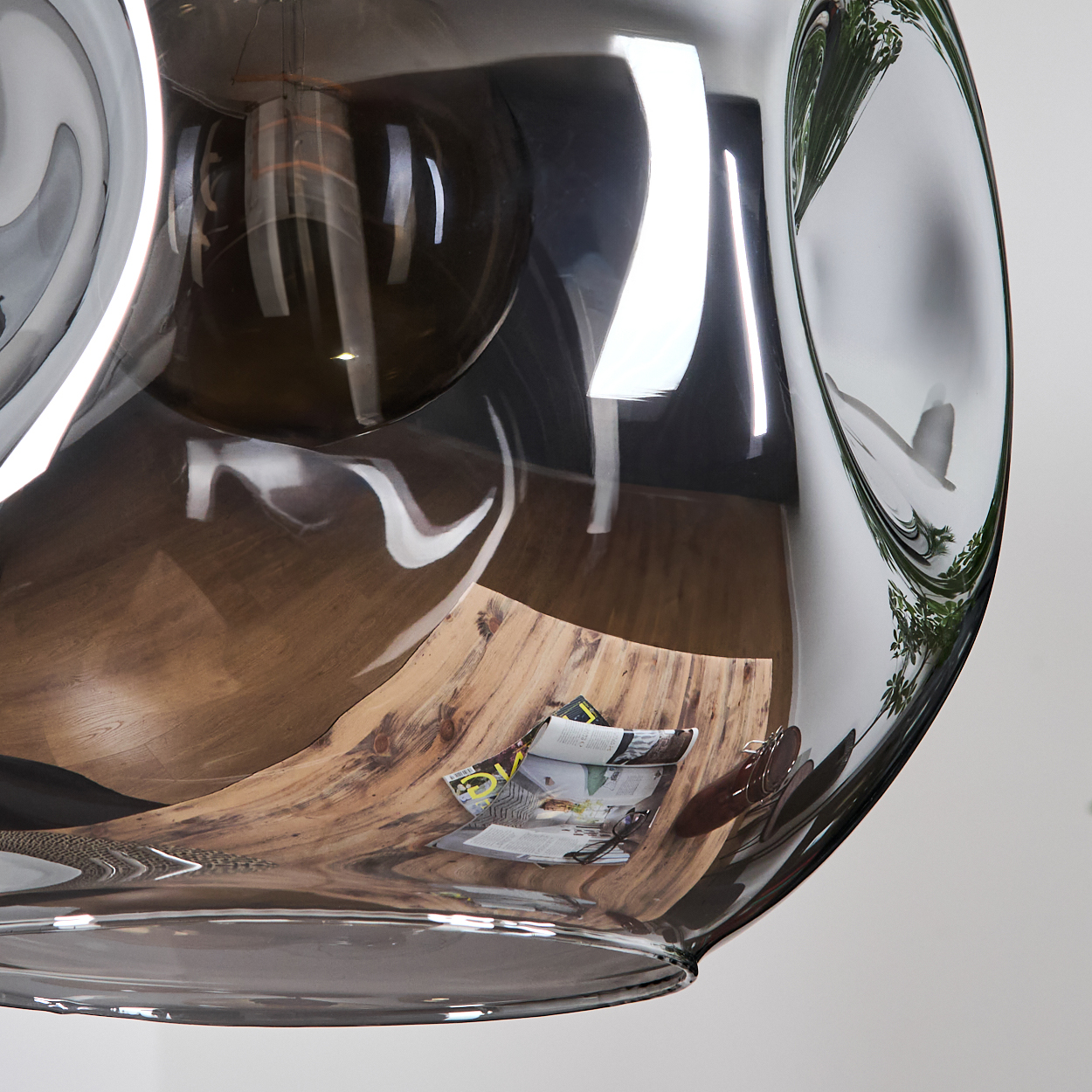 Ein verzerrtes, klares Glasobjekt reflektiert einen Holztisch mit Zeitschriften und eine weiße Wand mit Grün. Das Glas ist glänzend.