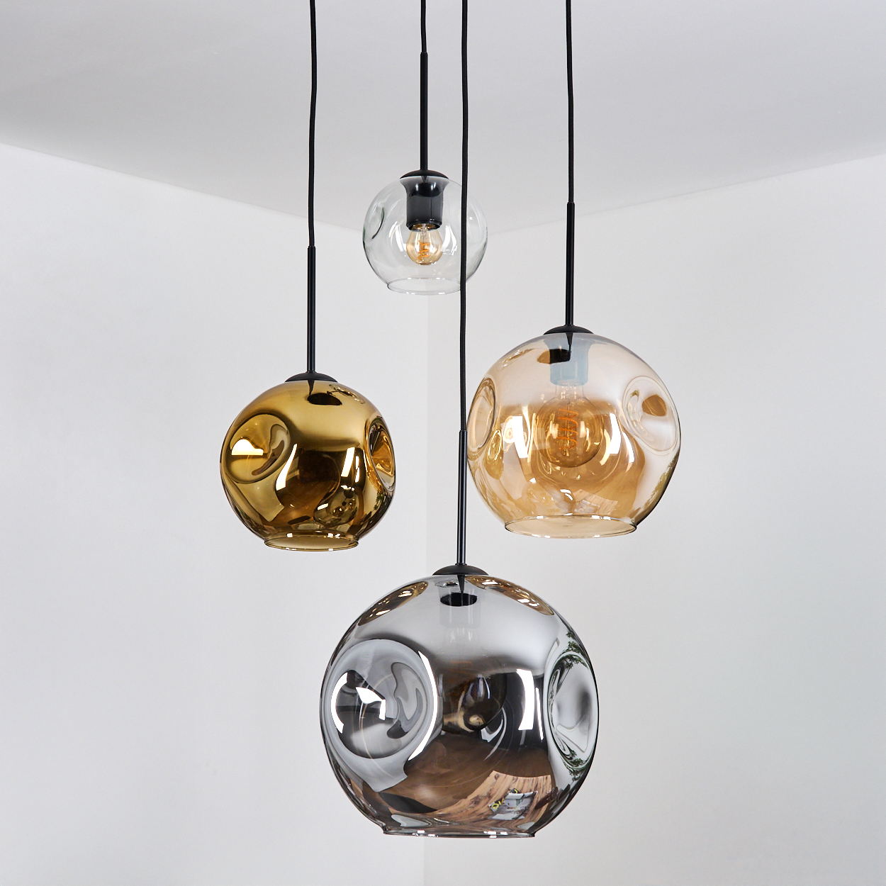 Vier moderne Pendelleuchten mit kugelförmigen Glasschirmen in Gold, Klar und Silber, die an schwarzen Schnüren vor einer weißen Wand hängen.