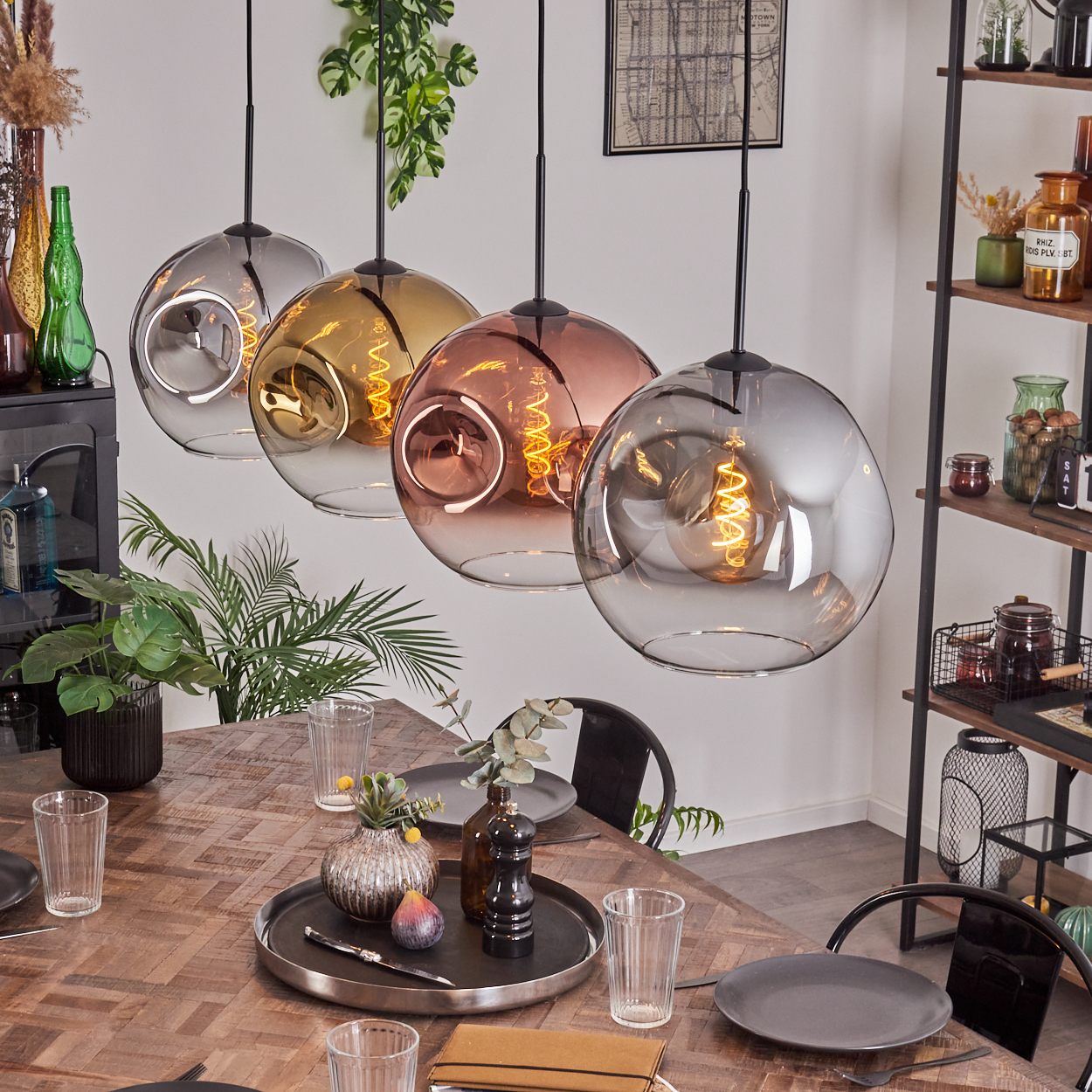 Vier bunte Glas-Pendelleuchten hängen über einem Holztisch. Der Tisch ist mit Tellern, Gläsern und einem Tablett mit Dekoration gedeckt.