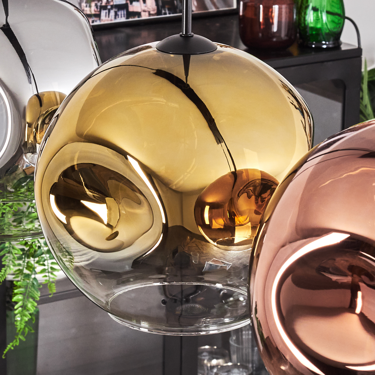 Nahaufnahme von drei kugelförmigen Glasschirmen in Gold, Silber und Kupfer. Der Hintergrund umfasst grüne Pflanzen und dunkle Möbel.