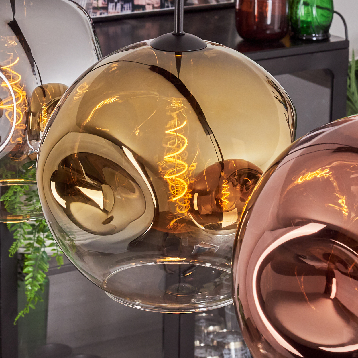 Nahaufnahme von drei modernen Pendelleuchten mit leuchtenden Wendelglühbirnen. Die Glasschirme sind bernsteinfarben, grau und bronze.