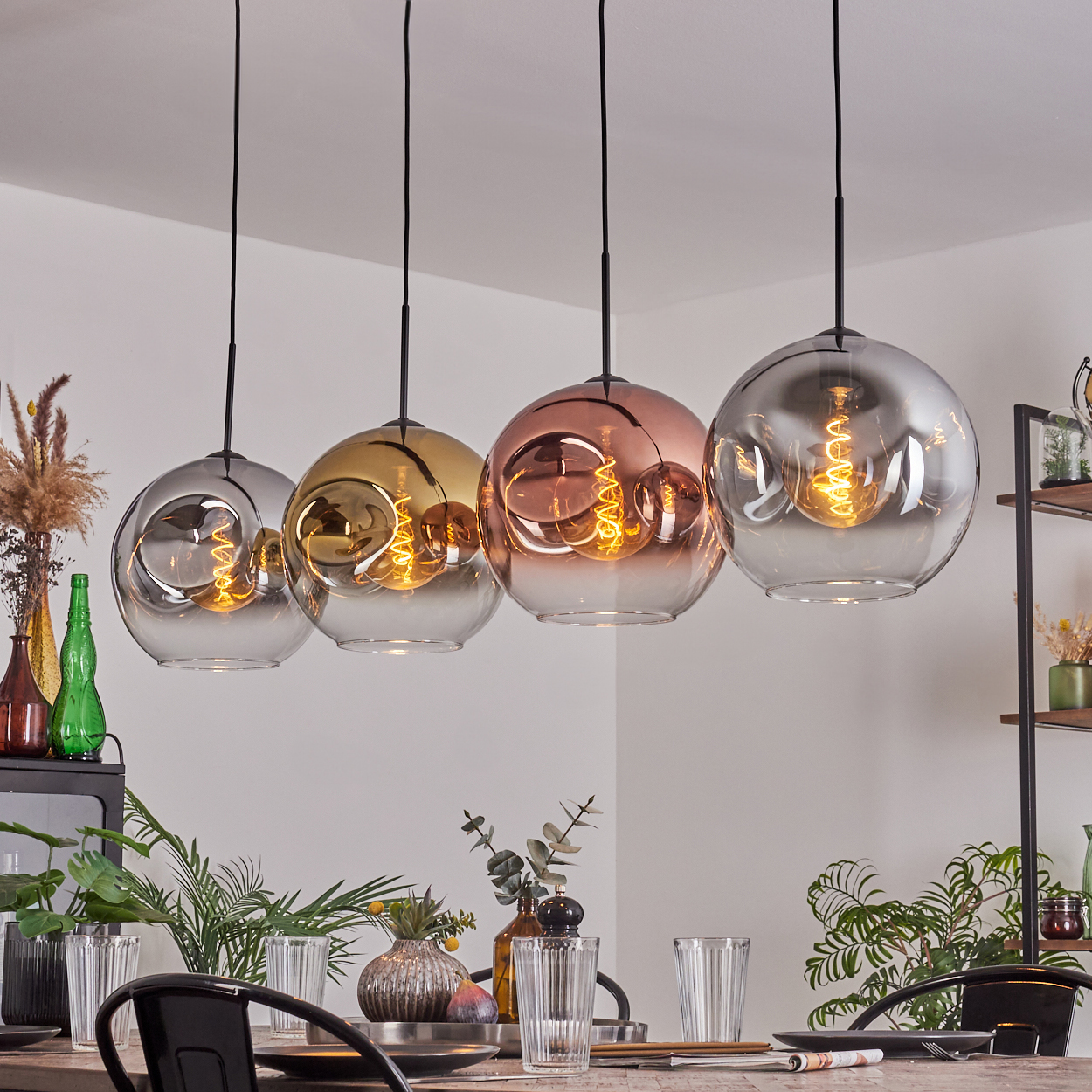 Vier Glas-Hängelampen in verschiedenen Farben hängen über einem Esstisch. Die Szene umfasst Pflanzen, Gläser und einen Holztisch.