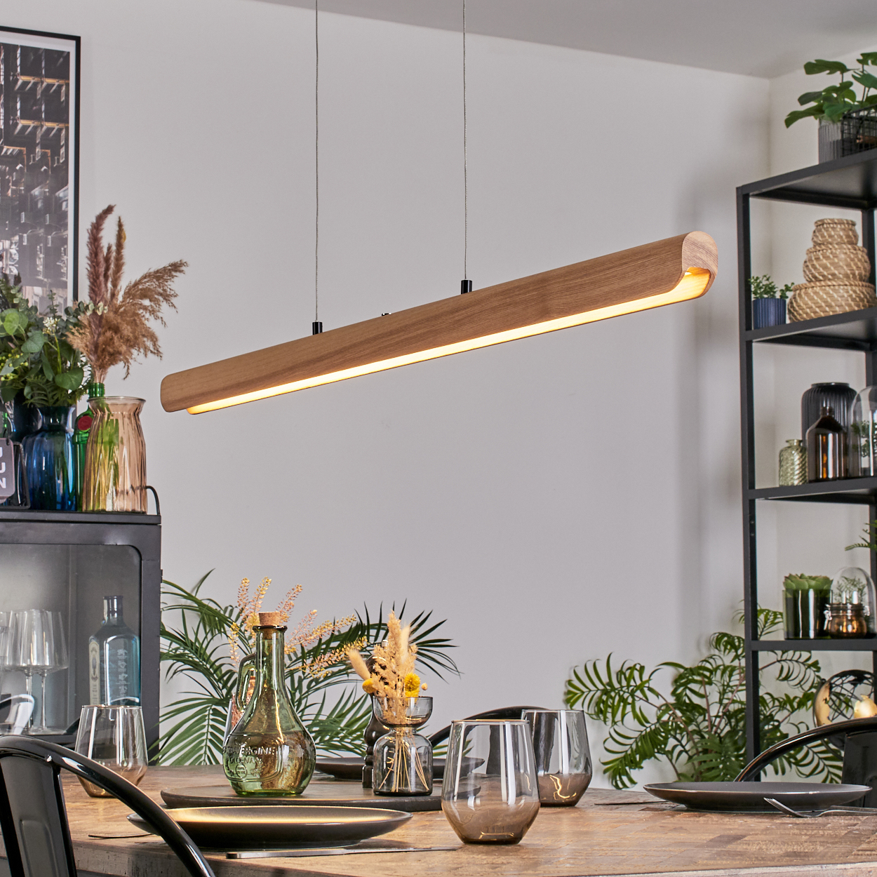 Ein Esszimmer mit Holztisch, beleuchtet von einer Holzleuchte. Vasen mit Blumen und Gläser stehen auf dem Tisch. Pflanzen und Regale sind sichtbar.
