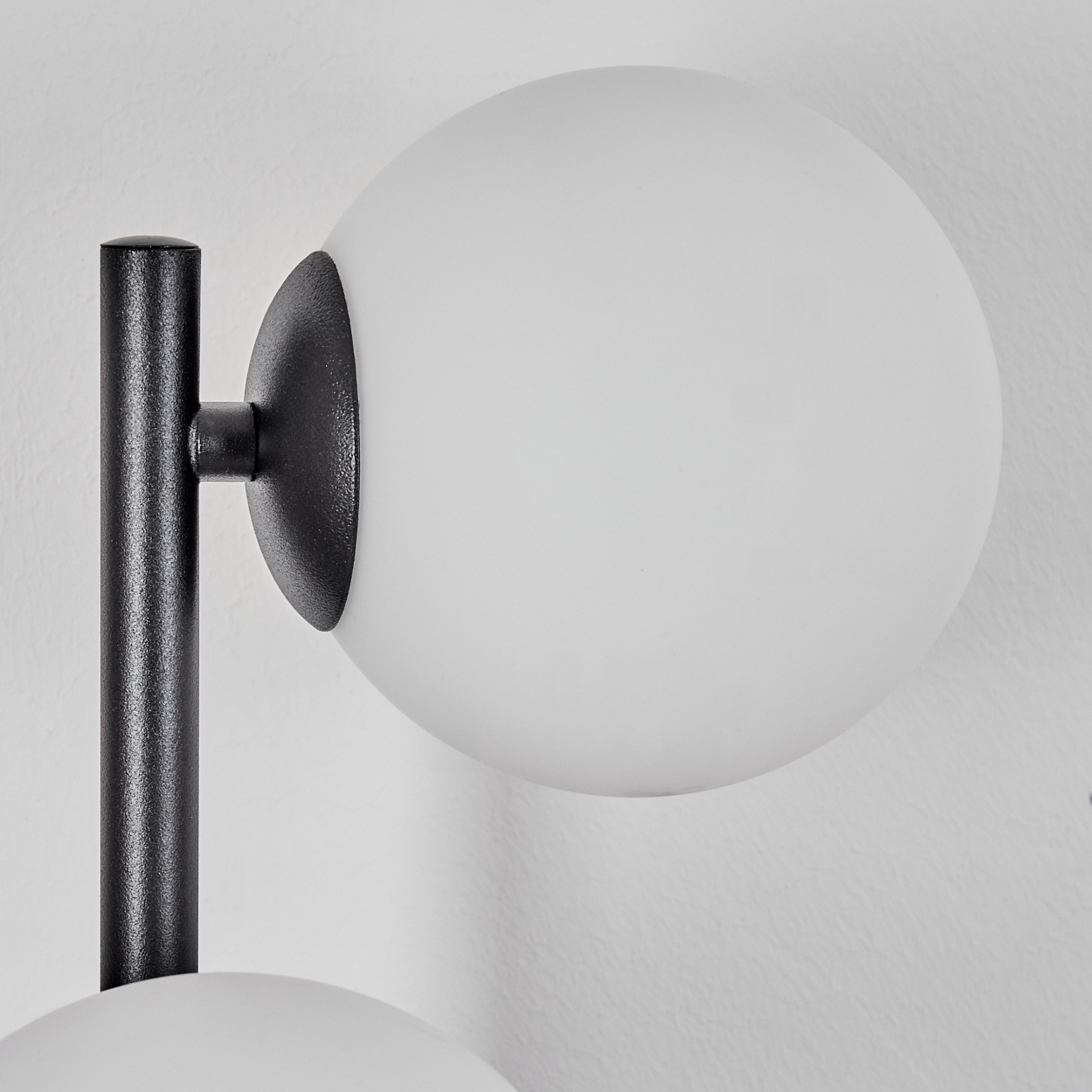 Eine moderne Lampe mit schwarzem Metallrahmen und zwei weißen, kugelförmigen Glasschirmen. Die Lampe ist an einer strukturierten weißen Wand befestigt.