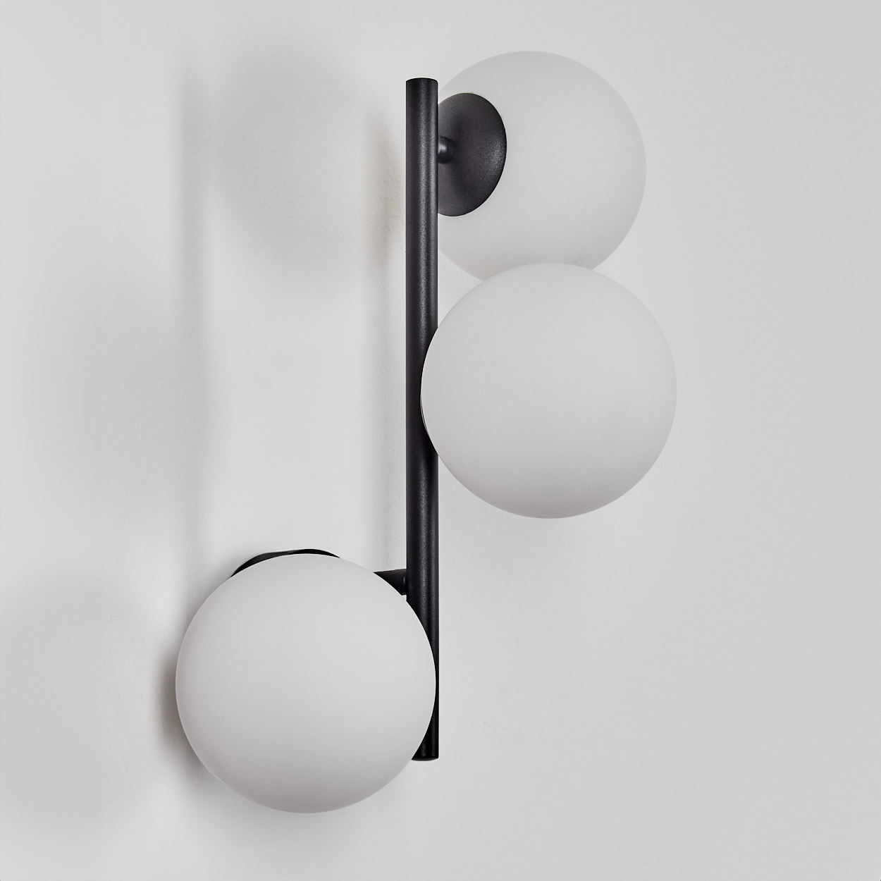 Eine moderne Wandlampe mit drei weißen, kugelförmigen Schirmen. Die Lampe hat ein schwarzes Metallgestell. Sie ist an einer weißen Wand montiert.