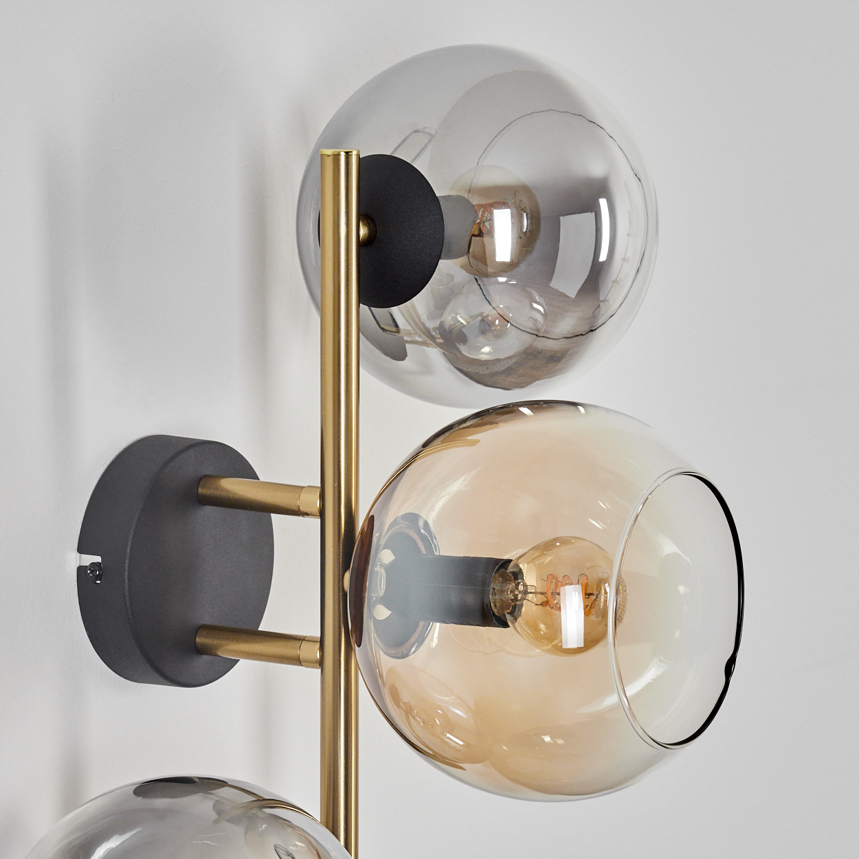Eine moderne Wandleuchte mit drei Glaskugeln. Kugeln sind klar, bernsteinfarben und grau. Die Lampe hat goldene und schwarze Metallakzente. An einer weißen Wand befestigt.