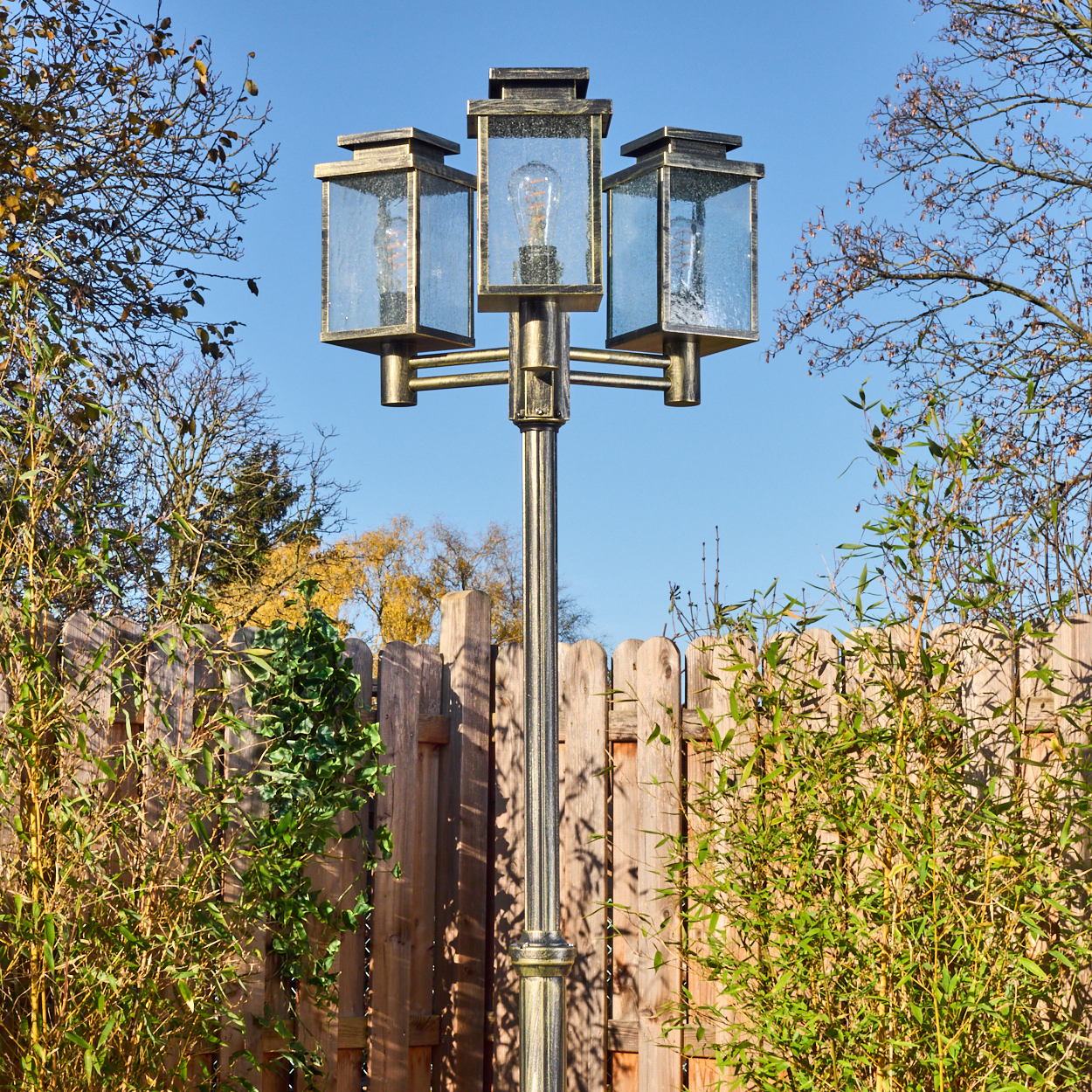 Eine dekorative Außenlampe mit drei Lichtern steht vor einem Holzzaun, umgeben von Bambus und Bäumen, vor blauem Himmel.